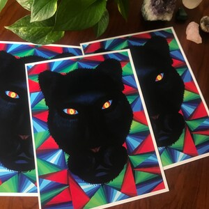 Może przedstawiać: Grafiki przedstawiające czarną panterę z uderzającymi żółtymi i pomarańczowymi oczami. Głowa pantery jest umieszczona na kolorowym geometrycznym tle z czerwonych, niebieskich i zielonych trójkątów. Grafiki są prezentowane na drewnianej powierzchni.