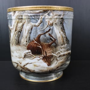May include: A cylindrical ceramic vase depicting a winter forest scene with deer. The vase has a gold rim and base, with a detailed painting of a snowy landscape and brown deer. The color scheme is muted with browns, whites, and blues.