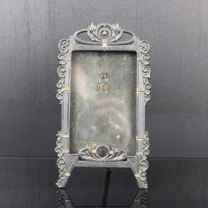 Puede incluir: Un marco de fotos plateado con un diseño ornamentado y un fondo negro. El marco tiene un soporte y está hecho de metal.