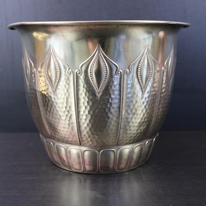 Pode incluir: Um vaso de metal prateado com textura martelada e detalhes verticais decorativos. O vaso tem uma borda larga e ligeiramente alargada e uma base arredondada com uma série de cristas verticais. Design clássico e elegante.