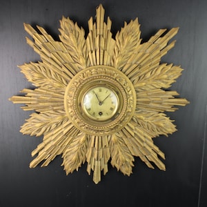 May include: A gold-colored sunburst wall clock with a round clock face. The clock has Roman numerals and ornate detailing. The sunburst design features radiating beams and leaf-like accents. The clock is mounted on a dark surface.