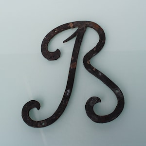 May include: A black metal letter R with a decorative, stylized design. The letter is made of wrought iron and has a rustic, aged appearance.