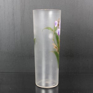 Pu&ograve; includere: Un vaso di vetro smerigliato di forma cilindrica. Il vaso &egrave; decorato con un motivo floreale con fiori viola, foglie verdi e dettagli dorati. Il vaso &egrave; vuoto e poggia su una superficie scura.