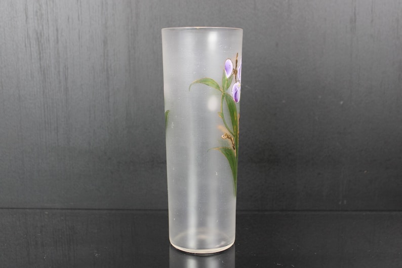 Pu&ograve; includere: Un vaso di vetro smerigliato di forma cilindrica. Il vaso &egrave; decorato con un motivo floreale con fiori viola, foglie verdi e dettagli dorati. Il vaso &egrave; vuoto e poggia su una superficie scura.