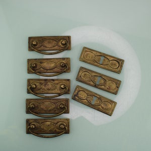 Puede incluir: Ocho tiradores de cajón de latón antiguo con diseños ornamentados. Los tiradores son rectangulares con un centro ovalado y una manija curva.