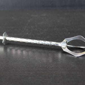 Peut inclure: Une pince à sucre en métal argenté dotée d'un long manche fin avec un motif à carreaux texturé. L'outil comprend un mécanisme à piston à ressort à une extrémité et une pince de préhension en forme de losange à cadre ouvert à l'autre.