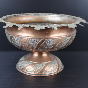 May include: A copper bowl with an ornate design and a pedestal base. The bowl has a decorative rim and is made of polished copper.