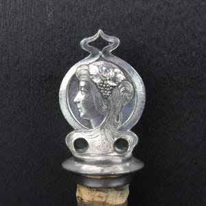 May include: A silver-colored bottle stopper with a detailed profile of a woman's face within a circular frame. The design includes floral and vine details, and the stopper is attached to a cork base.