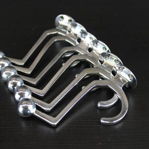 Puede incluir: Un conjunto de cinco ganchos de pared de metal plateado, cada uno con una base de montaje circular con dos orificios para tornillos, un brazo angular largo y un pomo esférico en el extremo. Los ganchos están dispuestos en pila.
