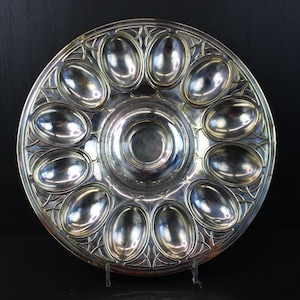 Peut inclure: Un plat de service argenté circulaire avec 12 compartiments ovales et un puits central. Le plat présente un design orné de style Art Déco sur le bord. Il est probablement utilisé pour servir des hors-d'œuvre, comme des huîtres.