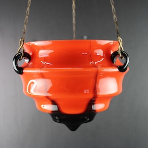 Op de afbeelding: Een hangende glazen kom met een feloranje lichaam en een getrapt, geribbeld ontwerp. De kom is voorzien van twee zwarte glazen handgrepen aan de zijkanten en een puntige zwarte glazen voet, opgehangen aan dunne metalen kettingen.