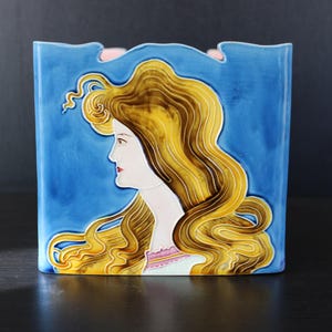 Puede incluir: Un jarrón cuadrado de cerámica que presenta el perfil en relieve de una persona con cabello dorado largo y ondulado sobre un fondo azul intenso. La persona tiene piel clara, labios rojos y viste una prenda clara con cuello ribeteado en rosa.