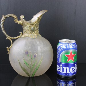 May include: A frosted glass jug with a silver-toned metal handle and spout. The jug has a floral design painted on the front and is sitting on a black surface next to a can of Heineken beer.