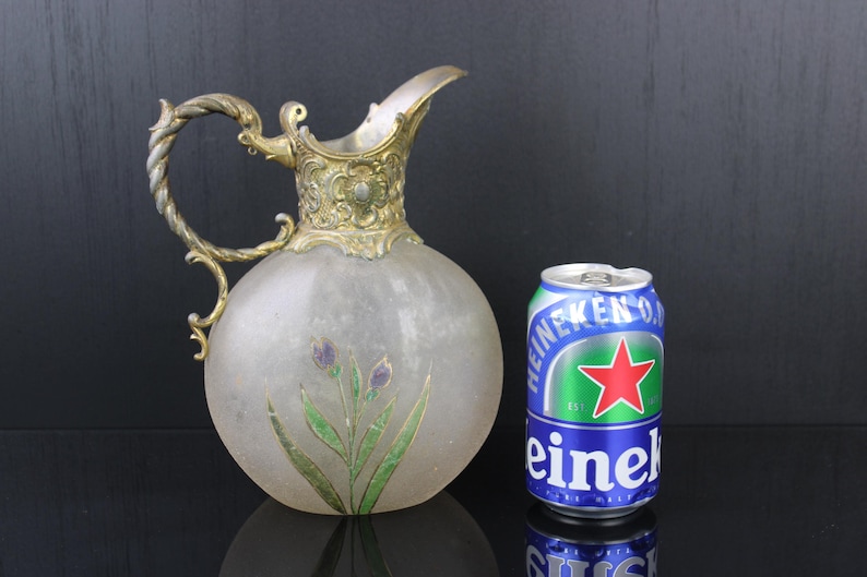 May include: A frosted glass jug with a silver-toned metal handle and spout. The jug has a floral design painted on the front and is sitting on a black surface next to a can of Heineken beer.