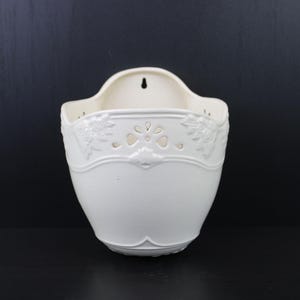 Puede incluir: Una maceta de pared de cerámica blanca con un diseño floral decorativo. La maceta tiene una parte superior curva con un agujero para colgar y una base redondeada. La parte delantera presenta un patrón en relieve detallado.