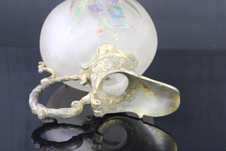 May include: A frosted glass jug with a gold metal handle and spout. The jug is decorated with a floral design in shades of blue, pink, and green.