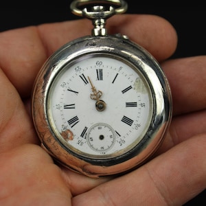 Puede incluir: Un reloj de bolsillo plateado con esfera blanca y números romanos negros. El reloj tiene un pequeño dial de segundos y una tija de cuerda plateada. El reloj se sostiene en una mano sobre un fondo negro.