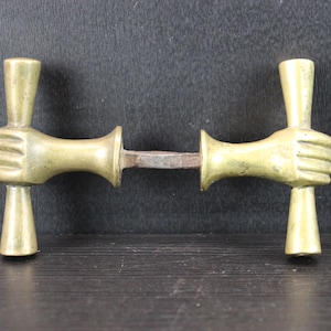 Könnte beinhalten: Ein Türgriffset aus Messing mit zwei symmetrischen Komponenten, die jeweils wie eine Hand geformt sind, die eine vertikale zylindrische Stange greift. Die beiden Teile sind durch einen zentralen quadratischen Metallstift verbunden und haben ein goldenes Finish.