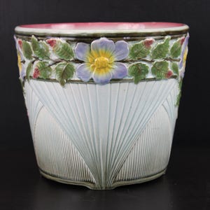 Op de afbeelding: Een keramische plantenpot met een witte basis en verticale ribbels. De bovenrand is versierd met een reliëfpatroon van groene bladeren, kleine rode bessen en paarse bloemen met gele harten. De binnenkant van de plantenpot is roze gekleurd.