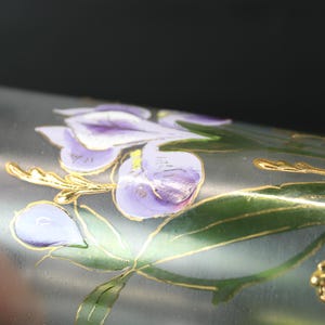 Pu&ograve; includere: Primo piano di un oggetto cilindrico argentato con un motivo floreale. Il motivo presenta fiori viola, foglie verdi e accenti dorati. L'oggetto sembra essere in metallo o vetro.