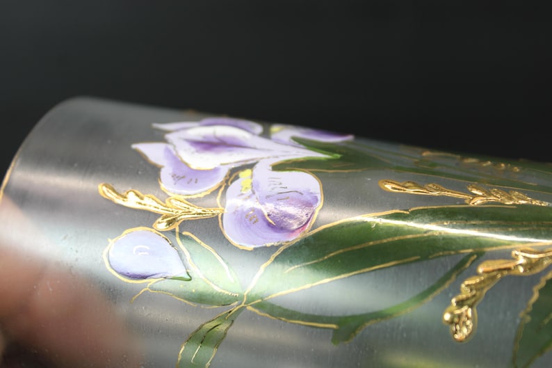 Pu&ograve; includere: Primo piano di un oggetto cilindrico argentato con un motivo floreale. Il motivo presenta fiori viola, foglie verdi e accenti dorati. L'oggetto sembra essere in metallo o vetro.
