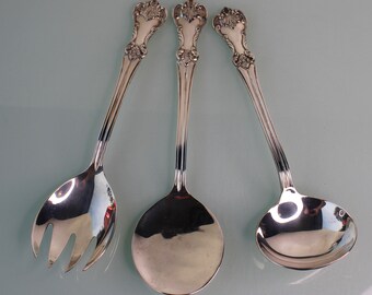 May include: Three silver serving spoons with ornate handles. The spoons are arranged in a triangular pattern on a light blue surface.