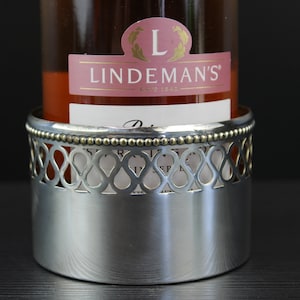 May include: A silver bottle holder with a decorative design and a beaded rim. The holder is holding a bottle of Lindeman's wine.
