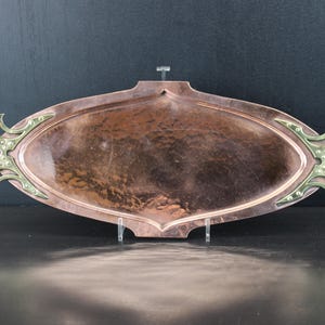 Pode incluir: Uma bandeja de servir oval e alongada em cobre com acabamento martelado e uma base central rebaixada. A bandeja apresenta duas alças ornamentadas em latão polido em cada lado, fixadas com rebites visíveis, criando um contraste distinto.