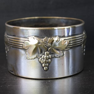 May include: Silver-toned metal wine coaster with a gold-colored grape and leaf design. The coaster has a cylindrical shape with a decorative band and is likely used for holding wine bottles.