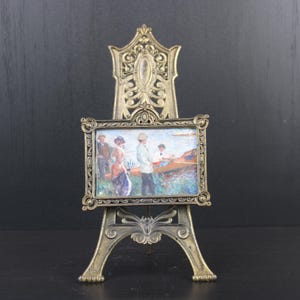 Op de afbeelding: Een gouden metalen ezel met een sierlijk ontwerp houdt een klein ingelijst schilderij vast van mensen in een boot. Het schilderij heeft een lichtblauwe achtergrond met een lichtblauwe lucht en water.