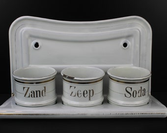 May include: White enamel wall-mounted storage rack with three white ceramic jars. The jars have gold trim and the words "Zand", "Zeep", and "Soda" written in gold lettering.
