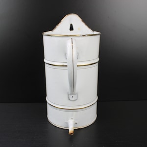 Puede incluir: Jarra de esmalte blanco con ribete dorado y asa. La jarra tiene un pico en la parte inferior y un agujero en la parte posterior para colgar.