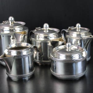 May include: A five-piece silver-toned tea and coffee service set featuring a tall coffee pot, a teapot, a smaller teapot, a creamer, and a lidded sugar bowl. Each piece has a polished metallic finish with a decorative beaded band around the base.