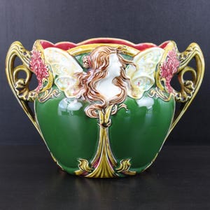 May include: A ceramic planter featuring a central relief of a figure with flowing hair and wings. The vessel is primarily deep green with gold and red accents, including ornate handles on both sides and a scalloped rim with a red interior finish.