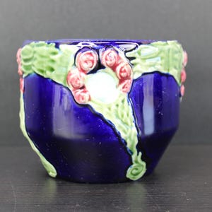 May include: A cobalt blue ceramic planter with a glossy finish. The rim is decorated with green leaves and pink roses. A white circular design is centered on the front. This decorative planter is suitable for small plants.