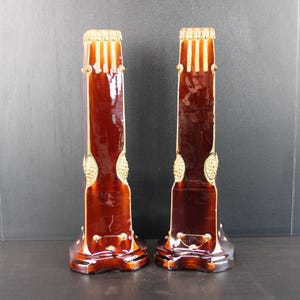 Pode incluir: Um par de castiçais de cerâmica castanha brilhante com um design canelado vertical no topo, detalhes dourados texturizados nas laterais e uma base alargada. Os castiçais têm um acabamento liso e refletor e uma forma estruturada.