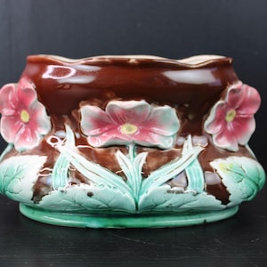 Puede incluir: Una maceta de cerámica marrón con acabado brillante, adornada con relieves de flores rosas y blancas y hojas verdes. La maceta tiene una parte superior ancha y abierta y una base redondeada, adecuada para uso en interiores o exteriores.