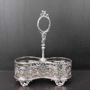 May include: Silver-plated condiment holder with an ornate floral design. The holder features two open compartments and a decorative handle with an oval opening. The base has decorative feet.