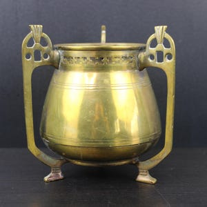 Könnte beinhalten: Eine antike Messingkanne mit Deckel, dekorativen Griffen und Füßen. Die Kanne hat einen abgerundeten Körper mit horizontalen Bändern und einen kleinen Griff auf dem Deckel. Das Messing hat eine warme, gealterte Patina.