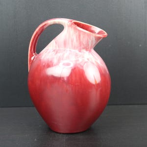 Peut inclure: Un pichet en céramique rouge et blanc avec un corps arrondi et une anse. Le pichet a une finition brillante et un motif tourbillonnant.