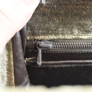 May include: Close-up of a brown leather bag with a zipper closure. The zipper is metal and has a pull tab with the word "LAKESTON" printed on it. The bag is lined with a light green fabric.