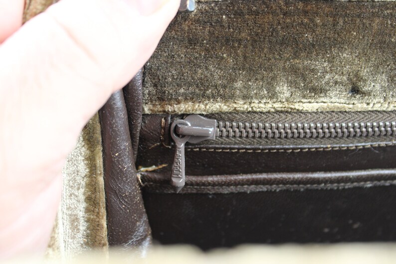 May include: Close-up of a brown leather bag with a zipper closure. The zipper is metal and has a pull tab with the word "LAKESTON" printed on it. The bag is lined with a light green fabric.