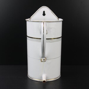 Puede incluir: Dispensador de agua de pared de esmalte blanco con ribete dorado y un pico en la parte inferior.