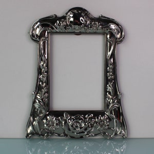 Puede incluir: Un marco de fotos plateado con un diseño ornamentado con motivos florales. El marco tiene una abertura rectangular para una foto o una obra de arte.