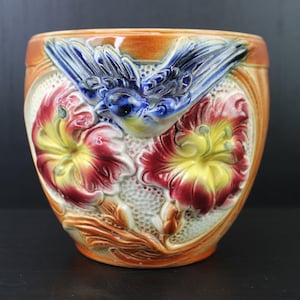 Puede incluir: Un jarrón de cerámica con un diseño en relieve detallado. El jarrón presenta un pájaro azul posado sobre dos vibrantes flores rojas y amarillas. El fondo tiene un patrón texturizado blanquecino, y el jarrón tiene una combinación de colores naranja y marrón.
