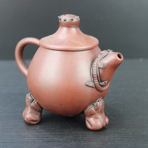 Op de afbeelding: Een terracotta theepot met een deksel en een tuit in de vorm van een draak. De theepot heeft drie poten in de vorm van kikkers.