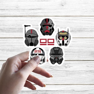 Pode incluir: Cinco adesivos de capacete de soldado clone Star Wars preto e cinza com detalhes vermelhos. Os adesivos estão sobre um fundo de madeira branca.