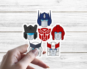 Könnte beinhalten: Ein Set mit sechs Transformers-Charakter-Kopfaufklebern. Die Aufkleber sind weiß mit schwarzen Umrissen und zeigen die Charaktere Optimus Prime, Megatron, Bumblebee, Starscream, Ironhide und Jazz. Die Aufkleber befinden sich auf einem weißen Holzgrund.