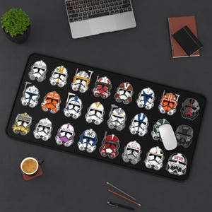 May include: A black mouse mat featuring a design of 28 different Star Wars clone trooper helmets. The helmets are in various colours, including red, blue, green, yellow, and orange. The mouse mat is rectangular and has a smooth surface.