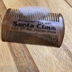 May include: A wooden comb with the text "Property of Santa Claus 123 Elf Rd. North Pole" engraved on it. The comb has fine teeth and is dark brown. A tuft of white material sits on top of the comb.